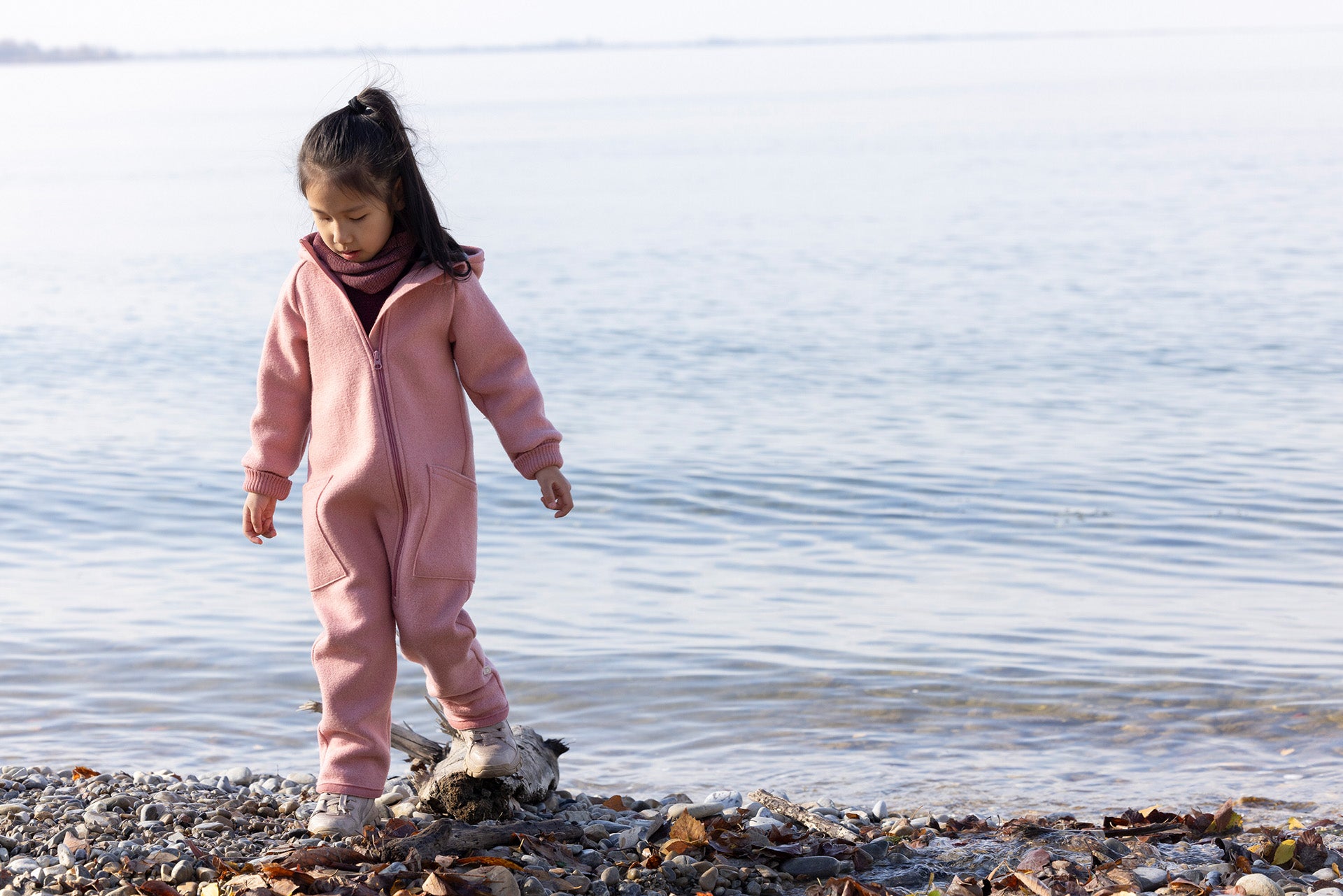 Child in a pink snowsuit standing on a rocky shore by a body of water.