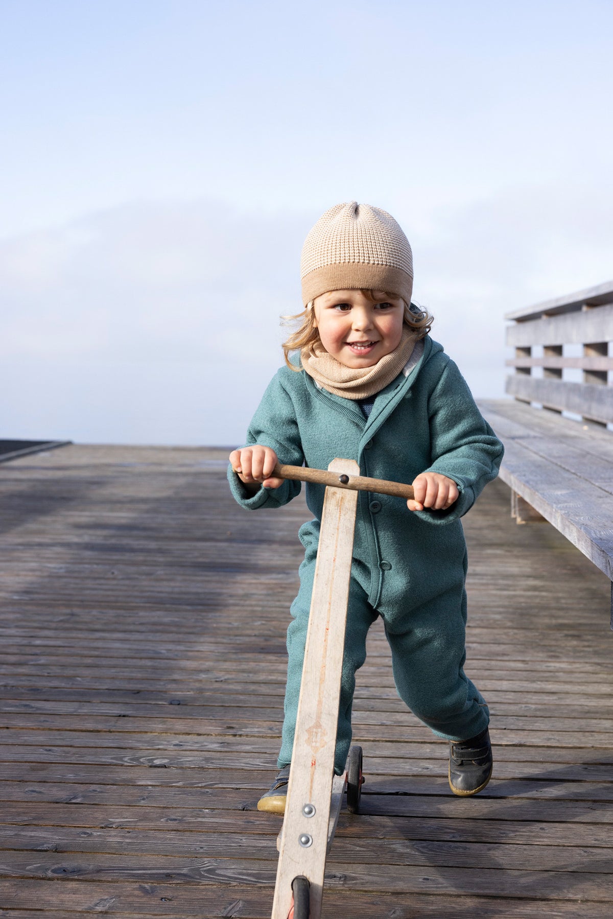 A toddler wearing a beige knit beanie, cream neck warmer, and a teal fleece one-piece outfit pushes a wooden toy scooter on a weathered wooden boardwalk.