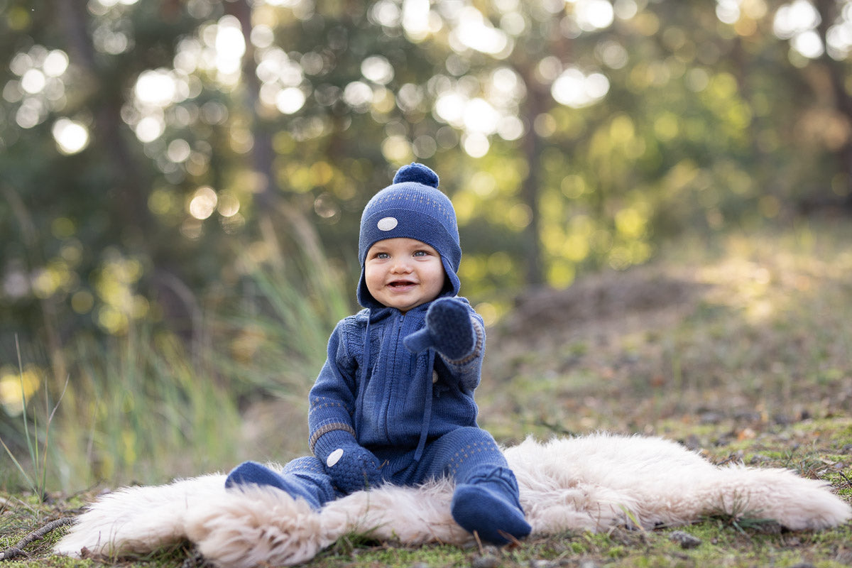 A baby wearing a navy blue knit hooded jumpsuit, matching hat, and mittens sits on a white faux fur rug outdoors in a wooded setting with a soft blurred background.