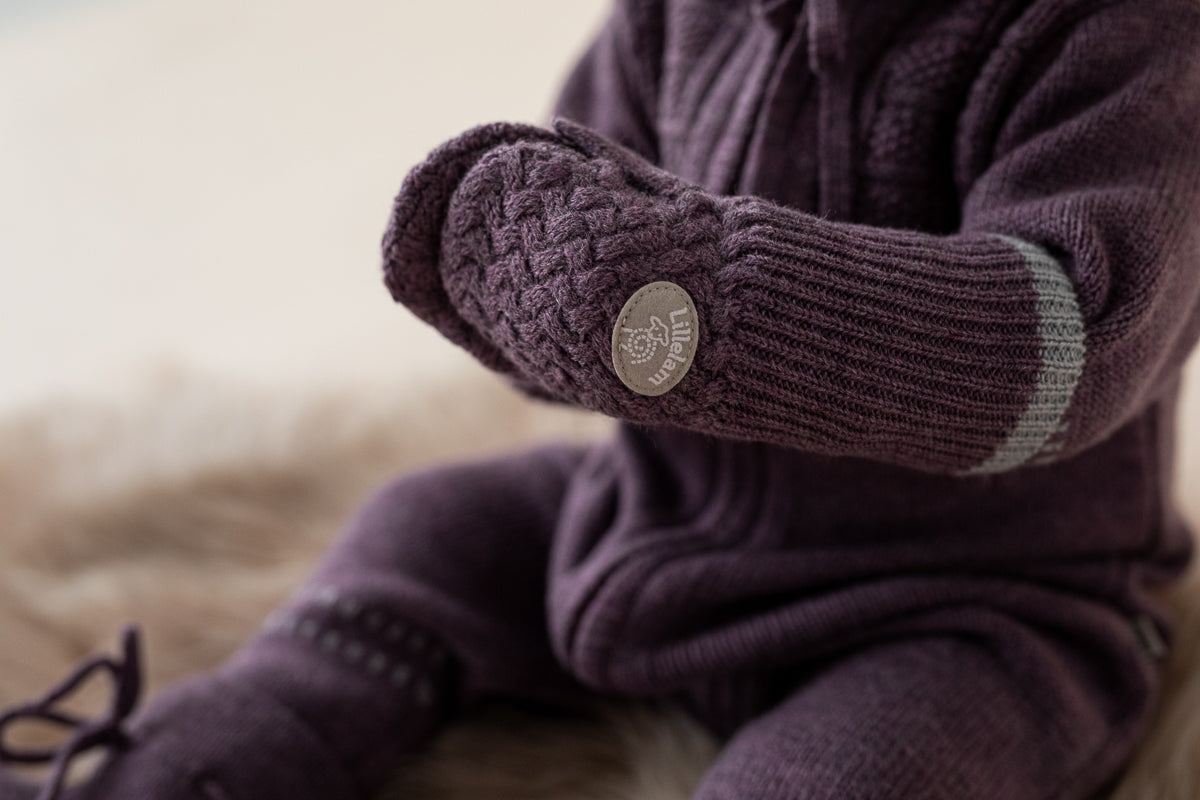 A close-up of a baby wearing a matching knit one-piece outfit and mittens in a deep plum purple color, sitting on a soft beige surface. The mittens feature a textured cable knit pattern, ribbed cuffs, and a round metal brand badge.