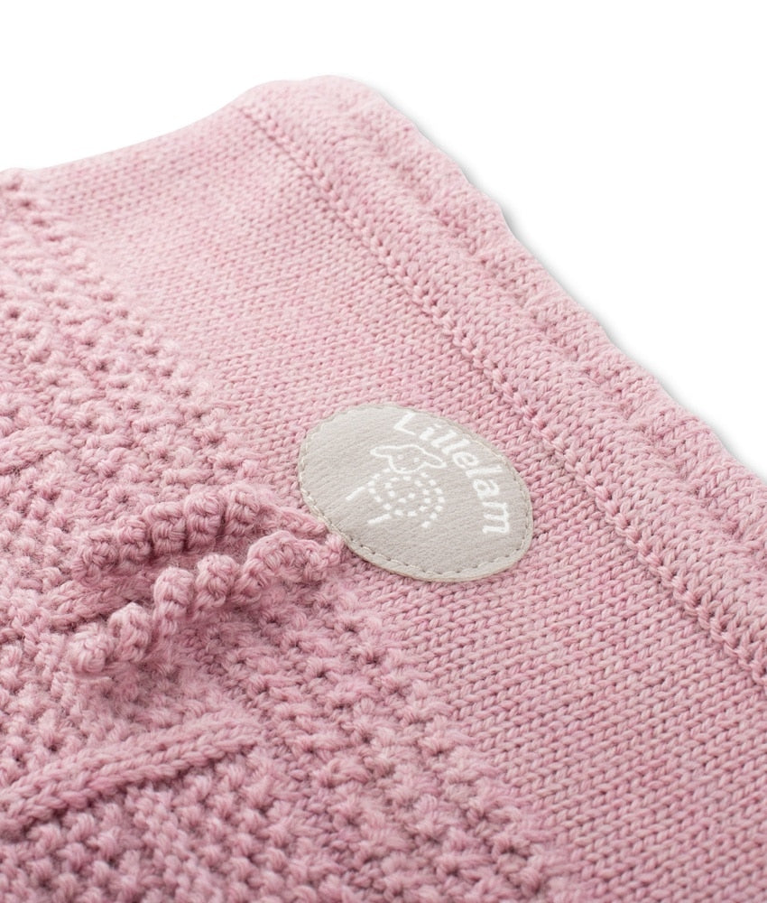 A close-up of a dusty pink knit textile, likely a baby blanket or swaddle, featuring a textured bobble pattern and a circular light gray brand patch reading 'Lil'elakm'.
