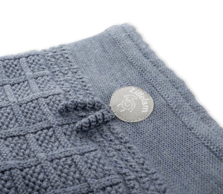 Close-up of a textured knit blanket in a dusty blue-gray color, featuring a small round white brand tag that reads 'Lil'lem' with a tree logo.