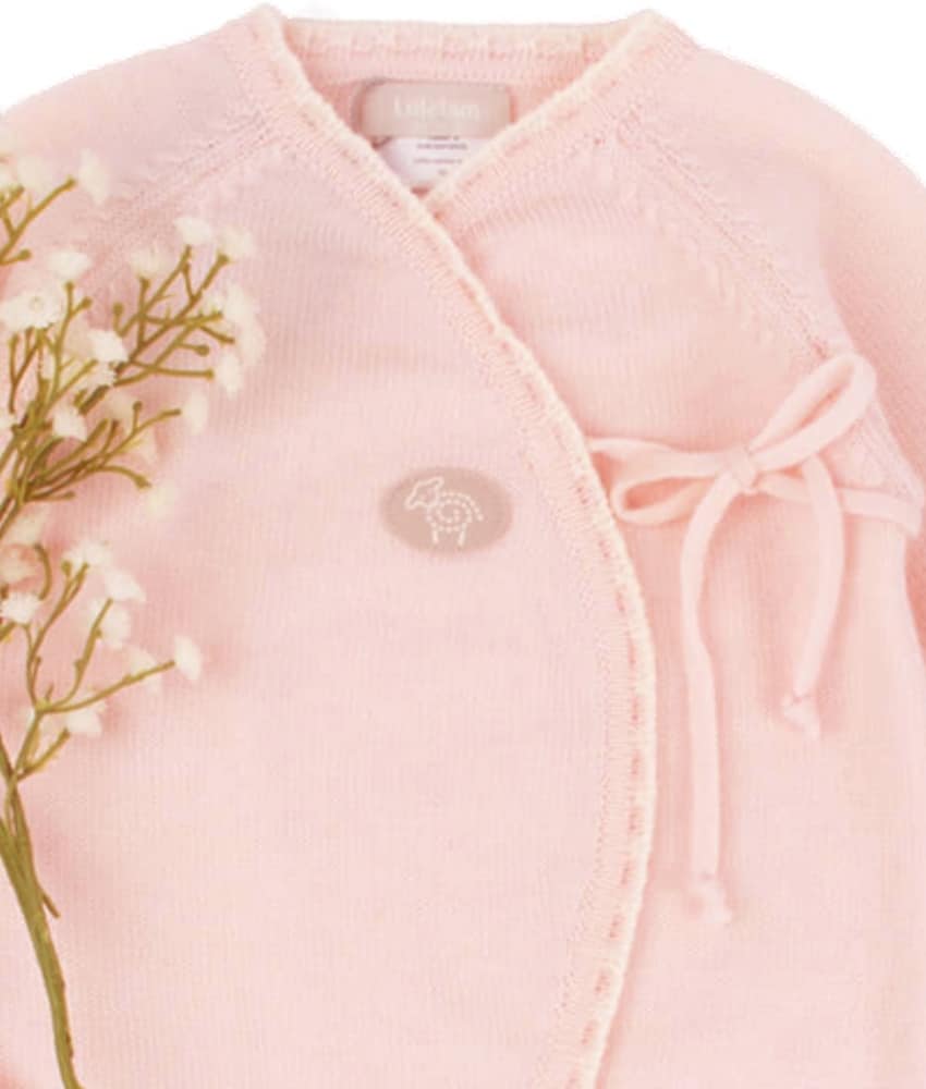 A close-up of a light pink knit baby wrap cardigan with a crossover front, ribbed trim, a drawstring tie on the chest, and a small embroidered logo patch. A floral stem with small white flowers is placed along the left side against a plain white background.