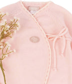 A close-up of a light pink knit baby wrap cardigan with a crossover front, ribbed trim, a drawstring tie on the chest, and a small embroidered logo patch. A floral stem with small white flowers is placed along the left side against a plain white background.