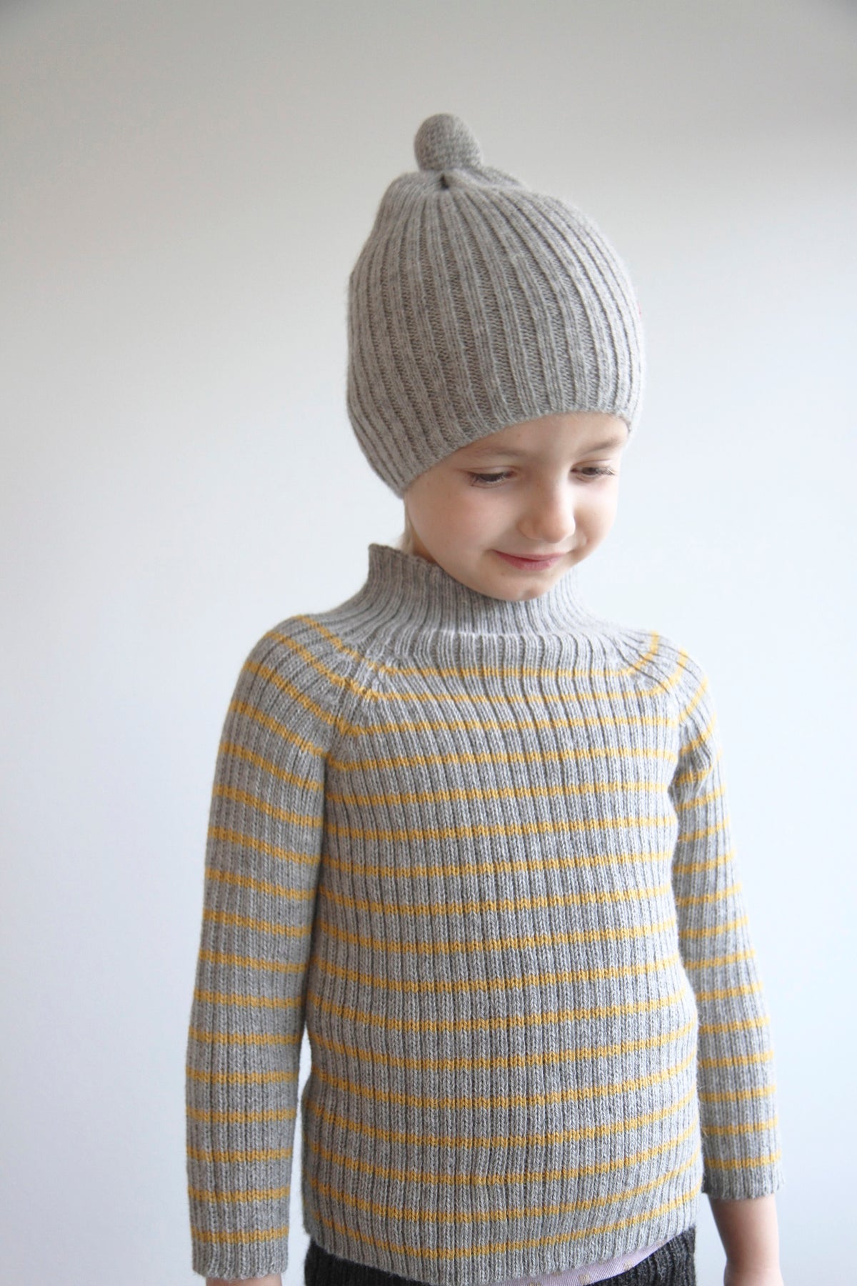 A child wearing a light gray ribbed knit beanie with a small pom-pom and a matching gray ribbed sweater with thin mustard yellow stripes, photographed against a plain light background.