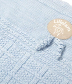 Close-up of a light blue knitted textile with a small oval beige Lilletam logo patch and knotted tassels.