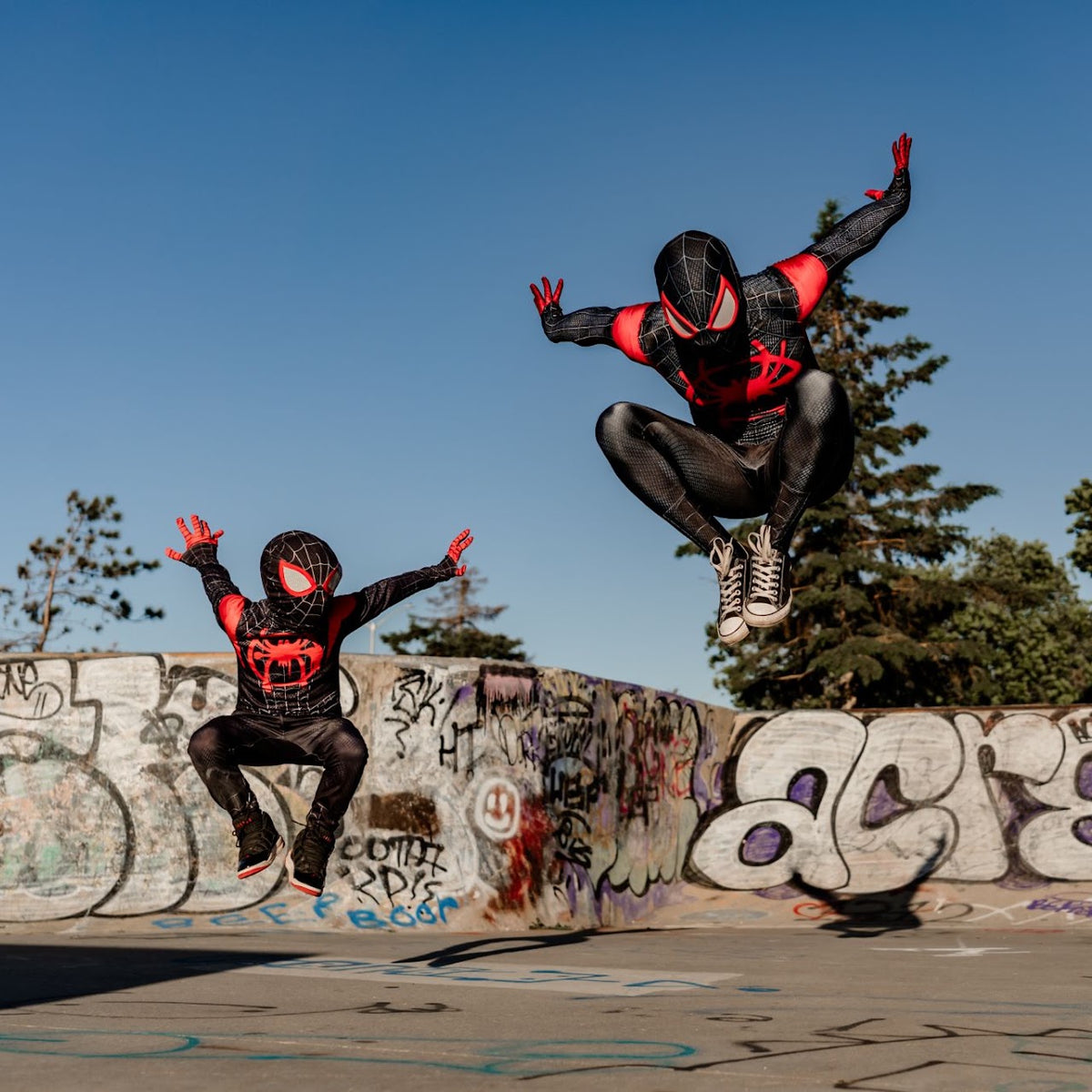 Two people wearing black and red Spider-Man style costumes jump in the air at an outdoor skatepark with graffiti walls and a blue sky background.